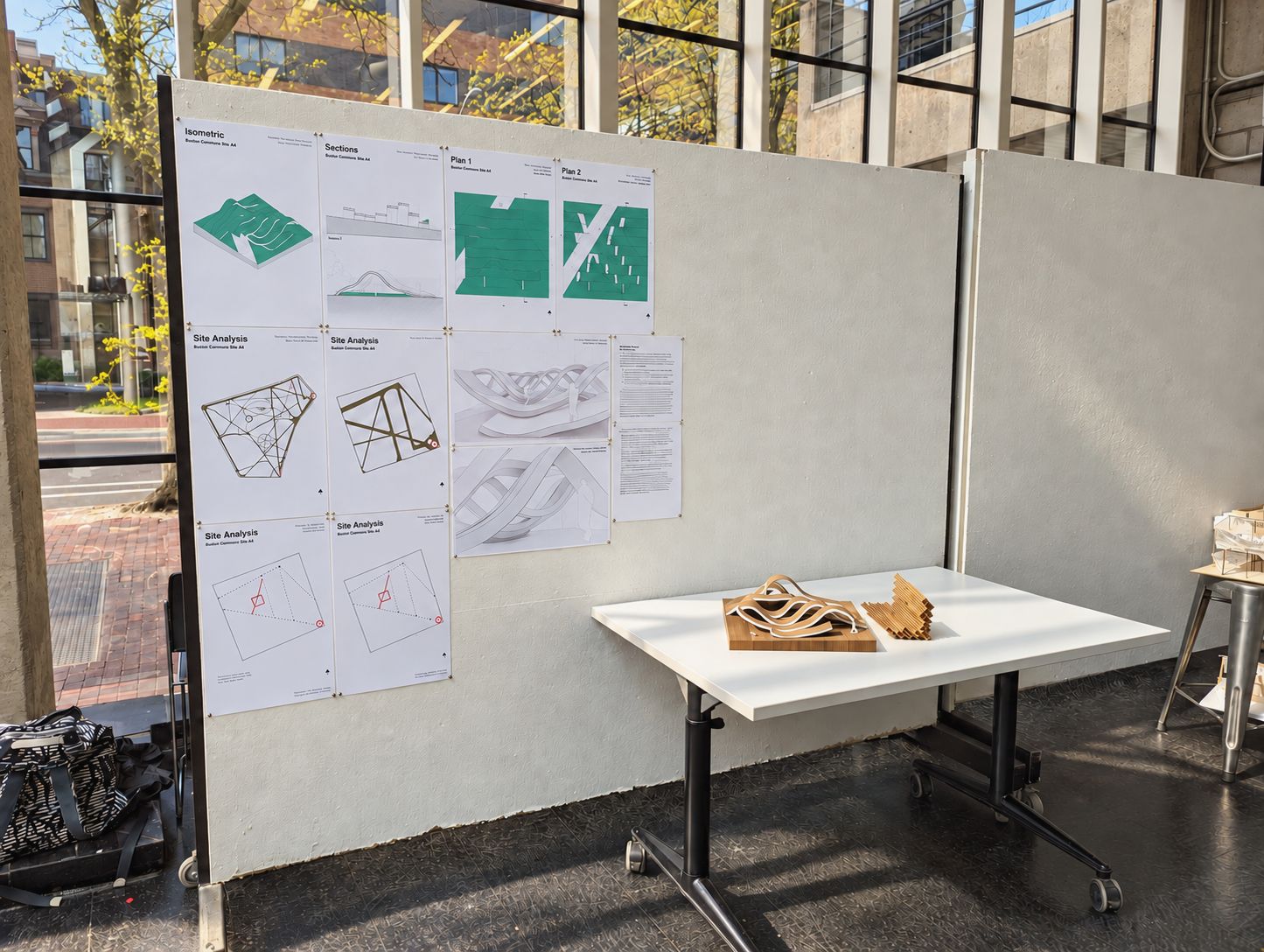 Architectural drawings pinned to a display board beside physical design models in a sunlit studio.
