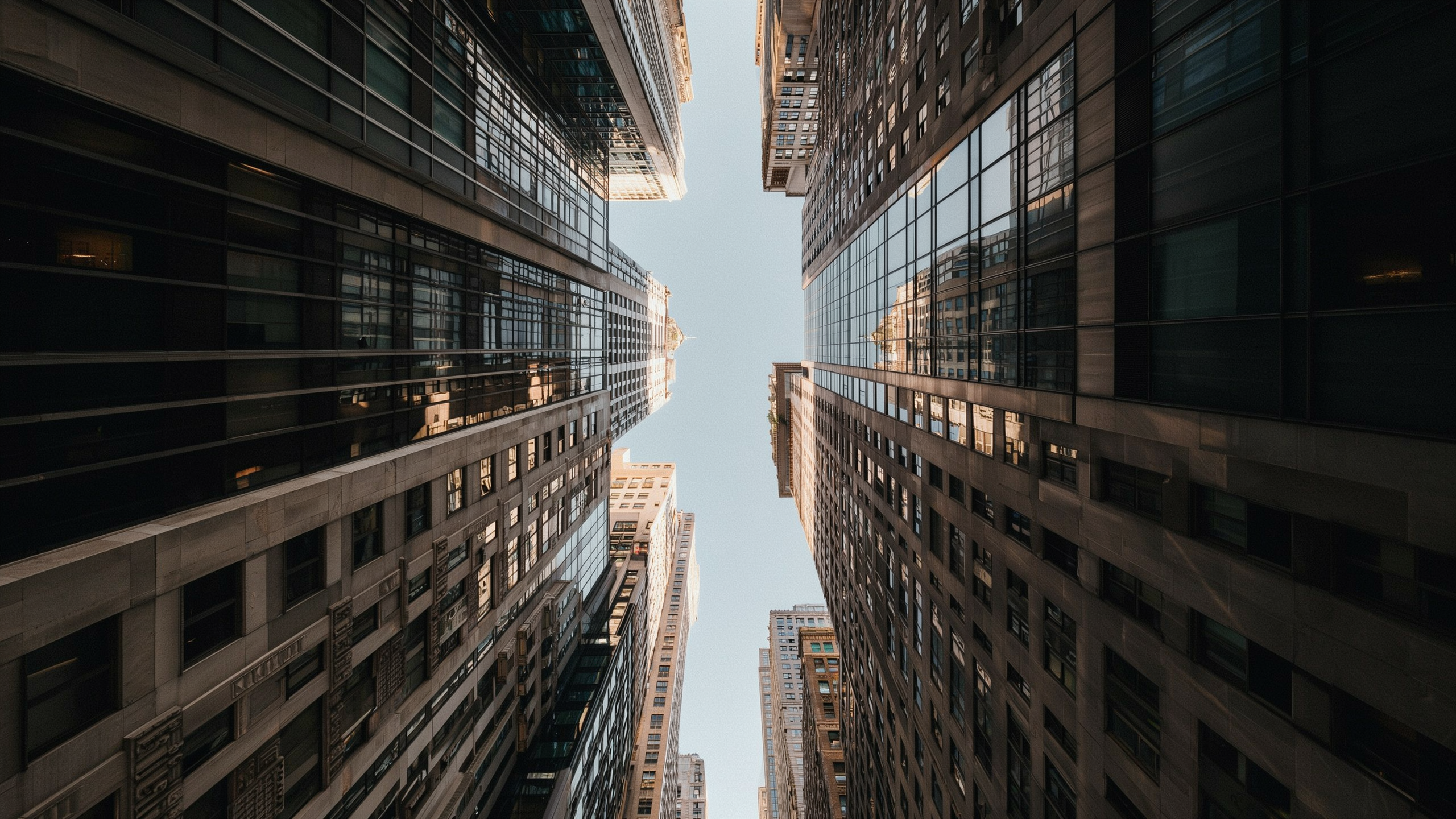 A cinematic upward view between stone and glass skyscrapers, with warm sunlight on upper facades and pale sky between the buildings.
